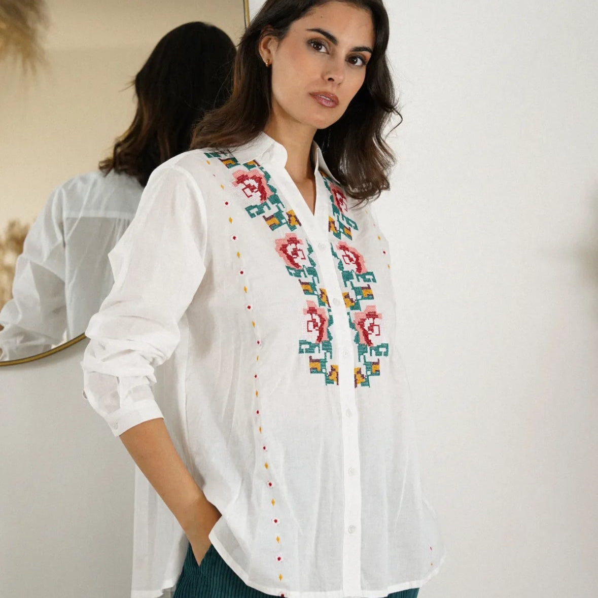 Woman wearing a white blouse with colorful embroidery standing in front of a mirror.