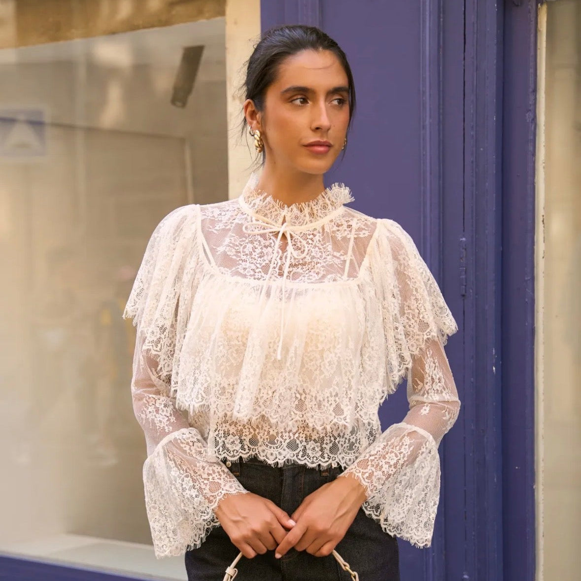 Model wearing KIDDING cream sheer lace blouse with high ruffle tie-neck, tiered chest overlay, and flared bell sleeves paired with dark denim jeans