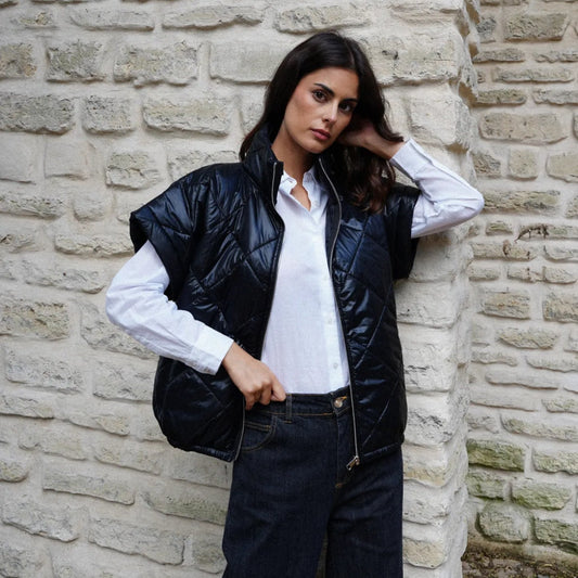 Woman wearing a black puffer vest over a white shirt against a stone wall.