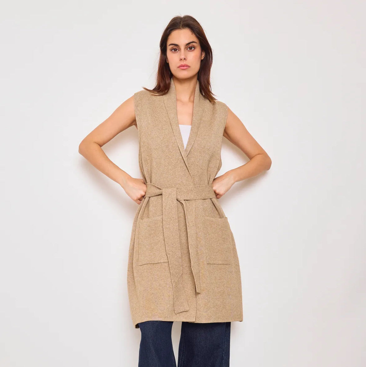 A female model wearing a sleeveless camel long gilet with a V-neckline and blue jeans.