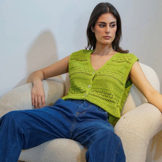 Woman wearing a green knitted vest and blue jeans sitting on a white chair.