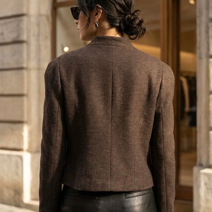 A woman viewed from behind wearing a cropped brown tweed blazer and black leather pants, standing in front of a stone building with an arched doorway.