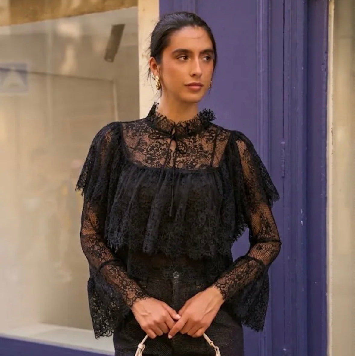 Model wearing KIDDING black sheer lace blouse with high ruffle tie-neck, tiered chest overlay, and flared bell sleeves paired with dark denim jeans"