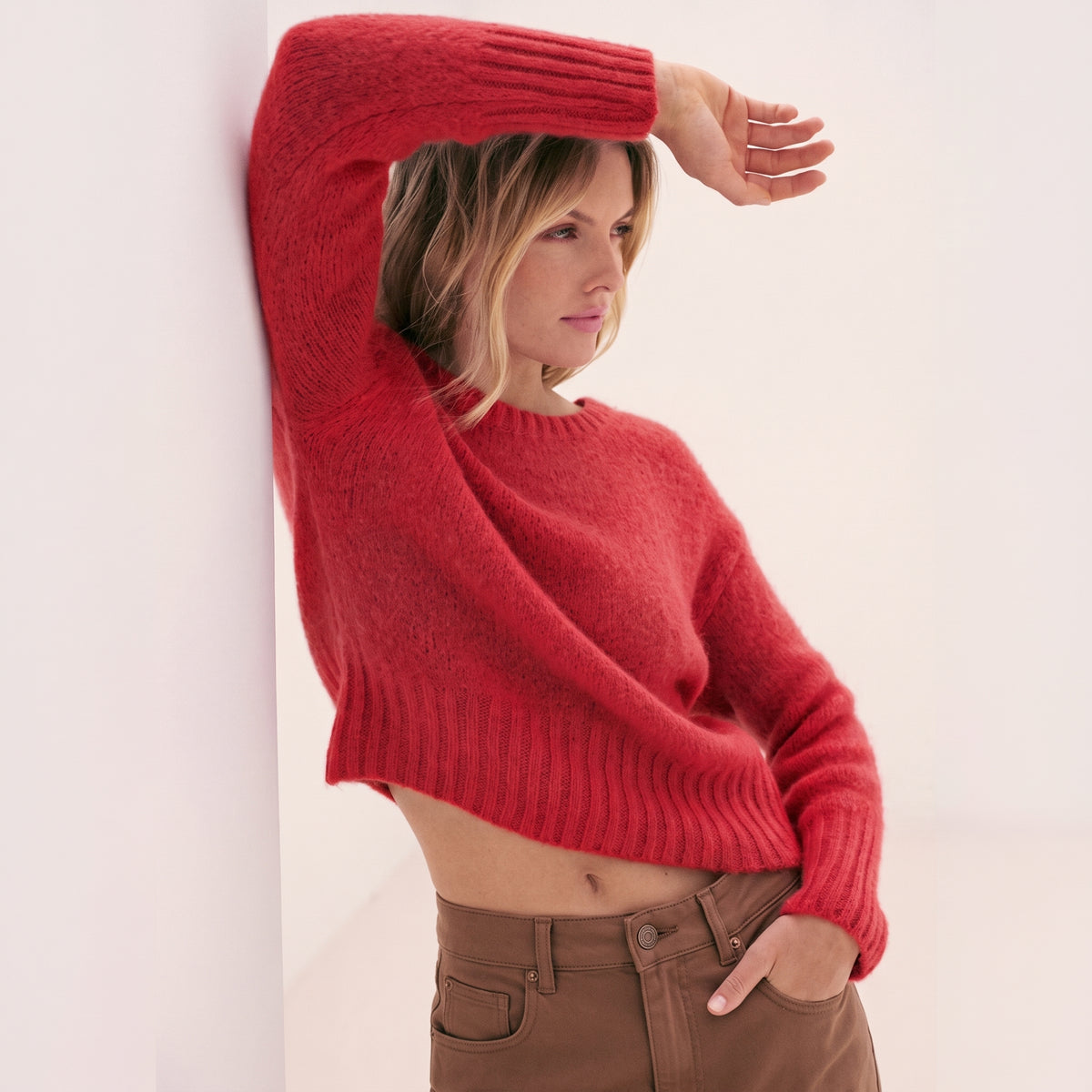 A blonde woman wearing a KIDDING bright red cropped plush jumper tucked into high-waisted brown trousers while leaning against a white architectural wall