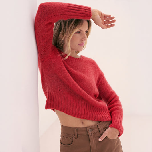 A blonde woman wearing a KIDDING bright red cropped plush jumper tucked into high-waisted brown trousers while leaning against a white architectural wall