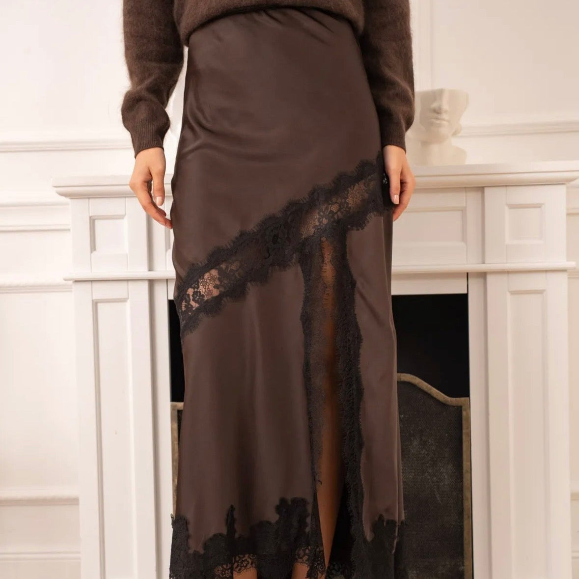 Model wearing KIDDING chocolate brown satin maxi skirt with diagonal black eyelash lace inset, high lace-trimmed leg slit, paired with an oversized brown sweater and black heels