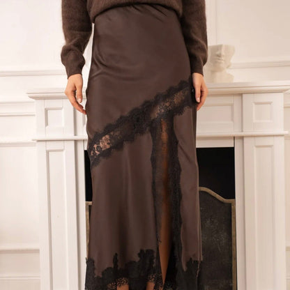 Model wearing KIDDING chocolate brown satin maxi skirt with diagonal black eyelash lace inset, high lace-trimmed leg slit, paired with an oversized brown sweater and black heels