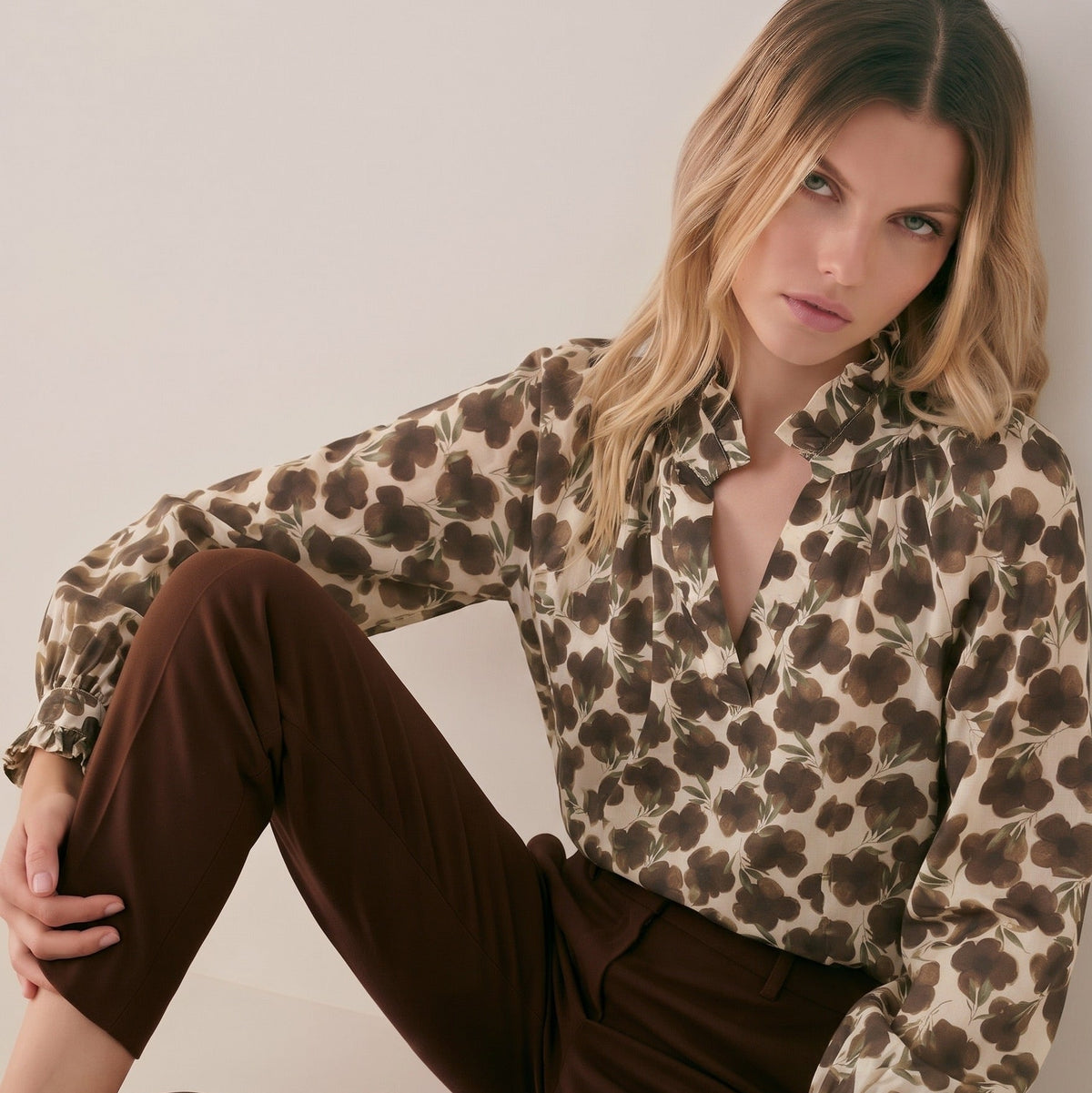 Woman seated against a neutral wall wearing a KIDDING long-sleeved cream blouse with a brown floral print and a ruffled neckline, paired with tailored dark brown trousers and classic brown leather loafers.