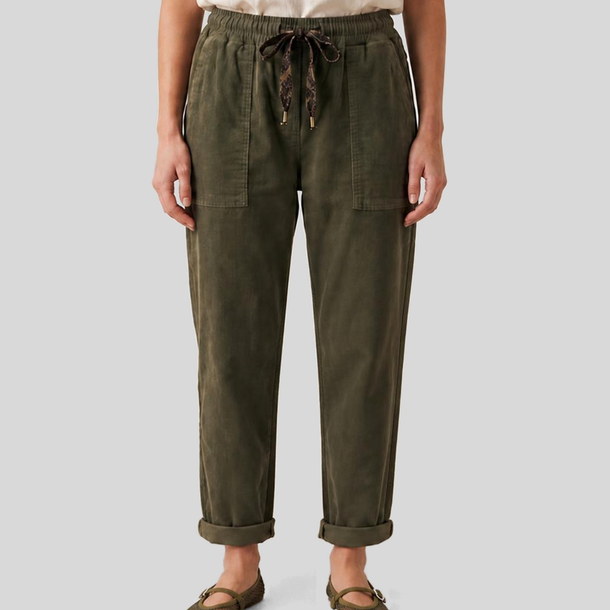 Front view from the waist down of a model wearing KIDDING olive green relaxed denim drawstring jeans with cuffed hems, paired with a cream t-shirt and olive green mesh Mary Jane flats.