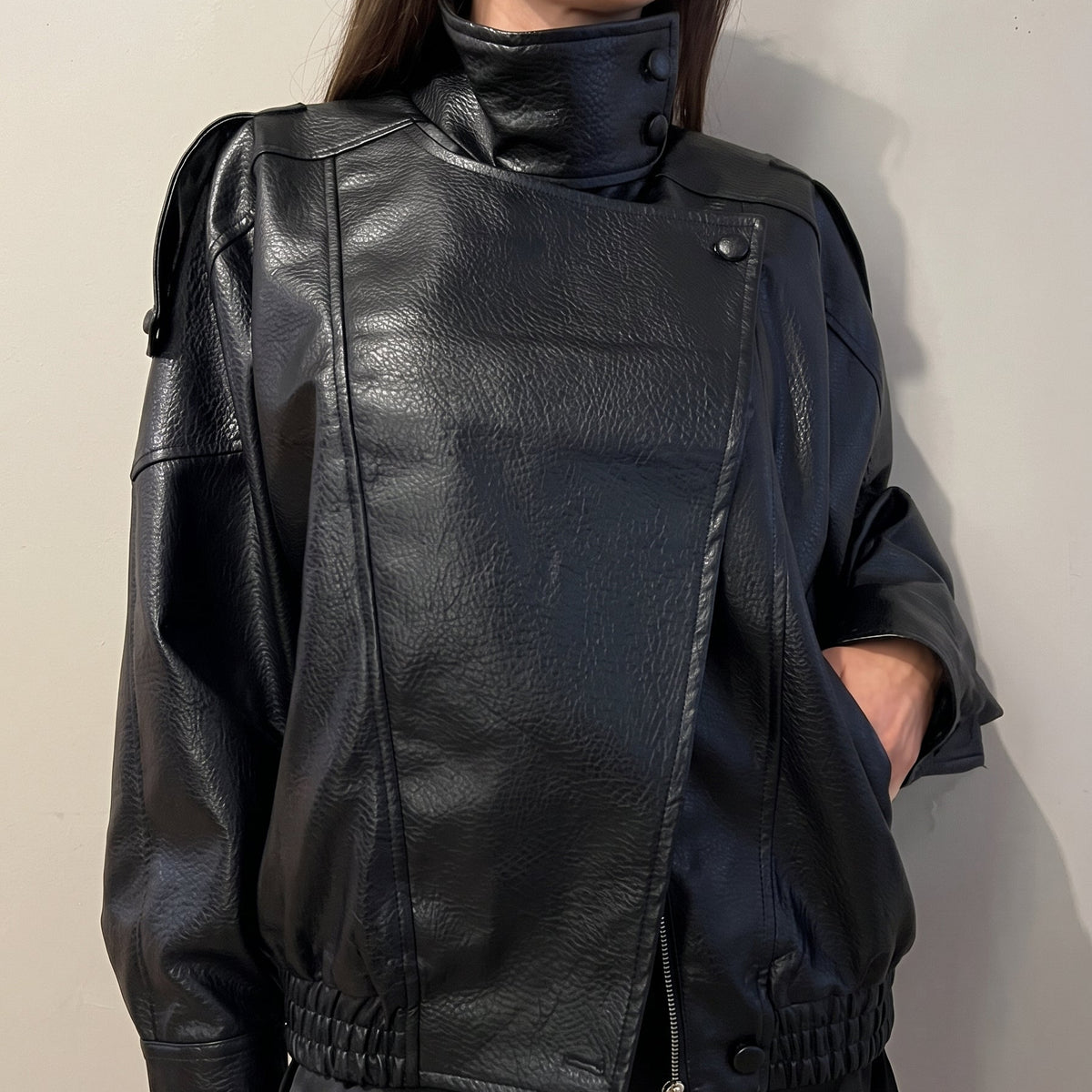 Close-up of a model wearing a kidding  oversized black pebbled faux leather jacket with a high stand collar, layered over a black lace-trimmed satin mini skirt.