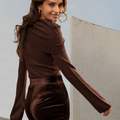 Smiling woman looking over her shoulder wearing a kidding rich chocolate brown long-sleeve knit top with a ribbed fold-over collar, styled with high-waisted dark brown velvet trousers featuring a satin waistband.